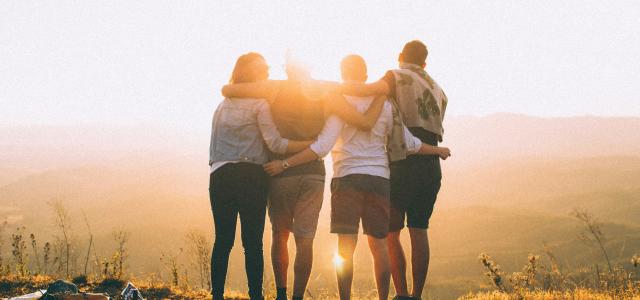 four person hands wrap around shoulders while looking at sunset by Helena Lopes courtesy of Unsplash.